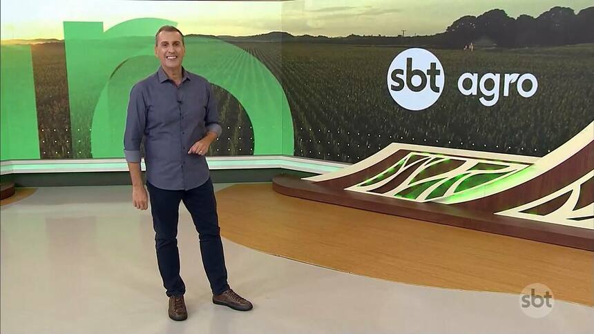 Sandro Ivanowski sorridente e em pé no centro do cenário do SBT Agro, ele vest calça preta e camisa azul