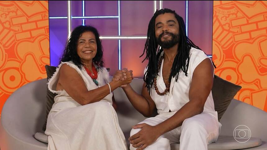 Ator Diogo Almeida e sua mãe Vilma, vestem branco, estão sorridentes e sentados no sofá do BBB 25