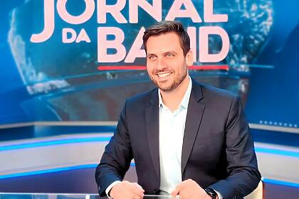 Douglas Santucci, sorridente, sentado na bancada do Jornal da Band