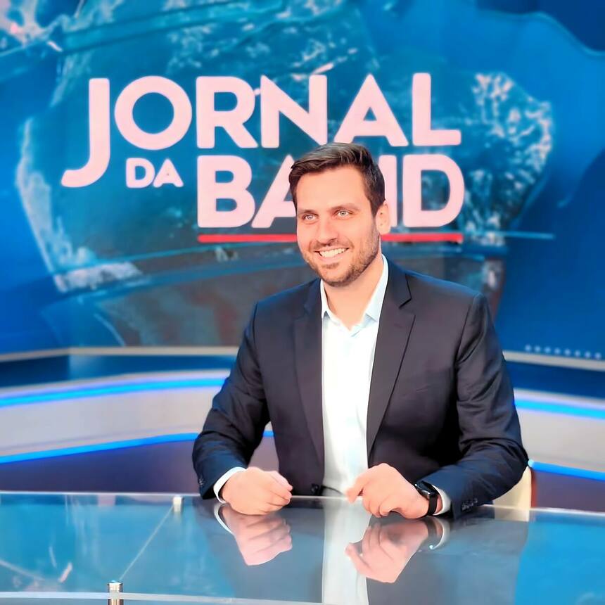 Douglas Santucci, sorridente, sentado na bancada do Jornal da Band