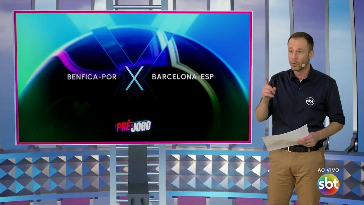 Tiago Leifert, com uniforme do SBT, em frente ao telão com o logo do Benfica e Barcelona no cenário da Champions League