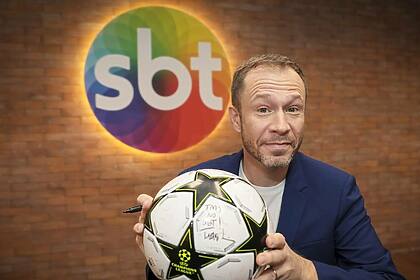 Tiago Leifert com uma bola nas mãos em frente ao logo do SBT