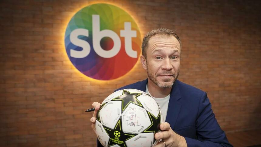 Tiago Leifert com uma bola nas mãos em frente ao logo do SBT