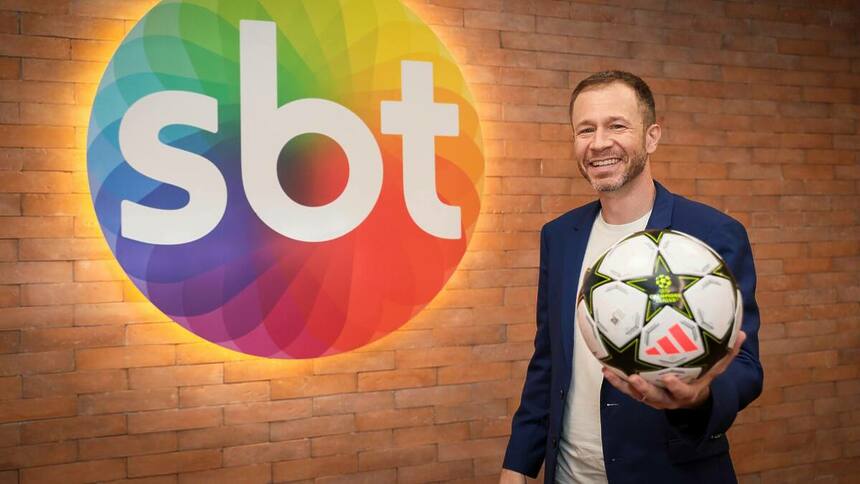 Tiago Leifert segurando uma bola e em frente ao logo SBT