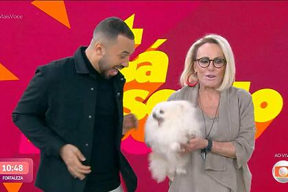 Gil do Vigor veste preto e está em frente ao telão do Mais Você com Ana Maria Braga segurando a cachorra que tentou morder o economista