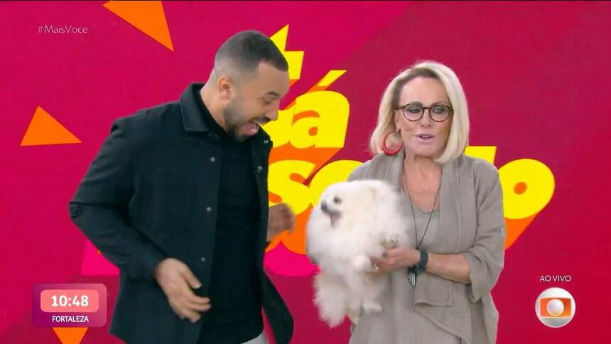 Gil do Vigor veste preto e está em frente ao telão do Mais Você com Ana Maria Braga segurando a cachorra que tentou morder o economista