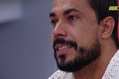 Maike chorando no Almoço do Anjo do BBB 25. Brother está com o olho cheio de lágrimas segurando uma almofada do patrocinador Claro