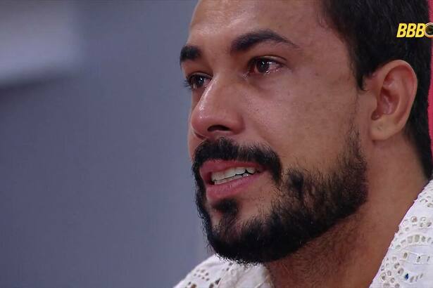 Maike chorando no Almoço do Anjo do BBB 25. Brother está com o olho cheio de lágrimas segurando uma almofada do patrocinador Claro