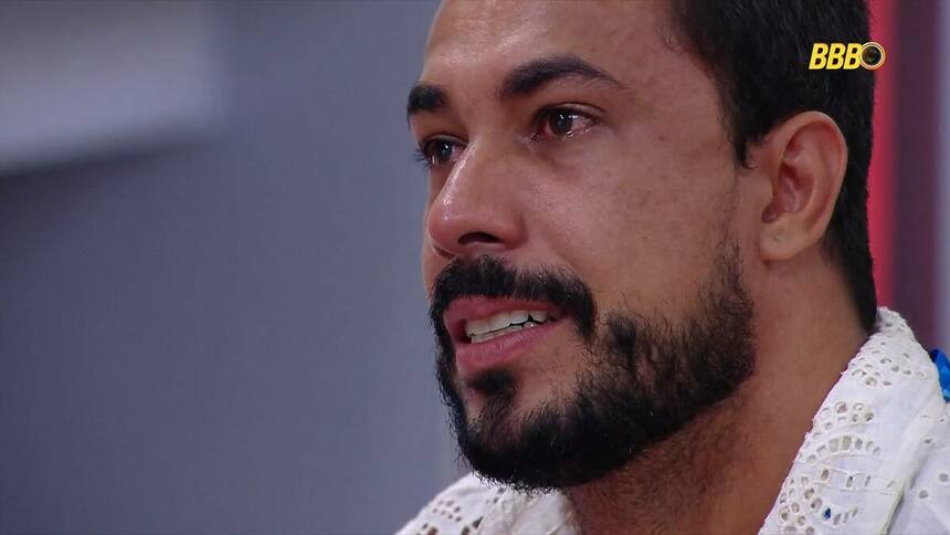 Maike chorando no Almoço do Anjo do BBB 25. Brother está com o olho cheio de lágrimas segurando uma almofada do patrocinador Claro