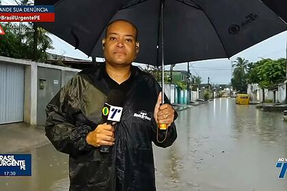 Apresentador Moab Augusto nas ruas de Recife, usando casaco de chuva e segurando um guarda-chuva e o microfone da TV Tribuna Band, apresentando o Brasil Urgente PE