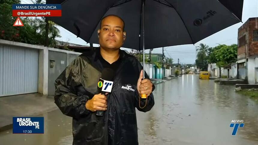 Apresentador Moab Augusto nas ruas de Recife, usando casaco de chuva e segurando um guarda-chuva e o microfone da TV Tribuna Band, apresentando o Brasil Urgente PE