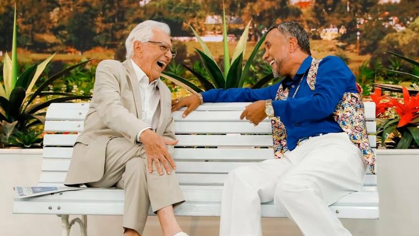 Carlos Alberto de Nóbrega e Paulinho Gogó sorridentes, de frente a frente, no banco da A Praça é Nossa II