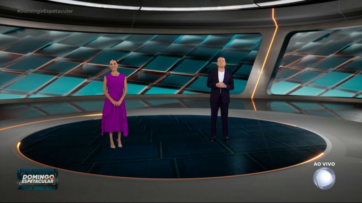 Roberto Cabrini e Carolina Ferraz no centro do cenário do Domingo Espetacular. Ele veste terno preto e ela conjunto lilás