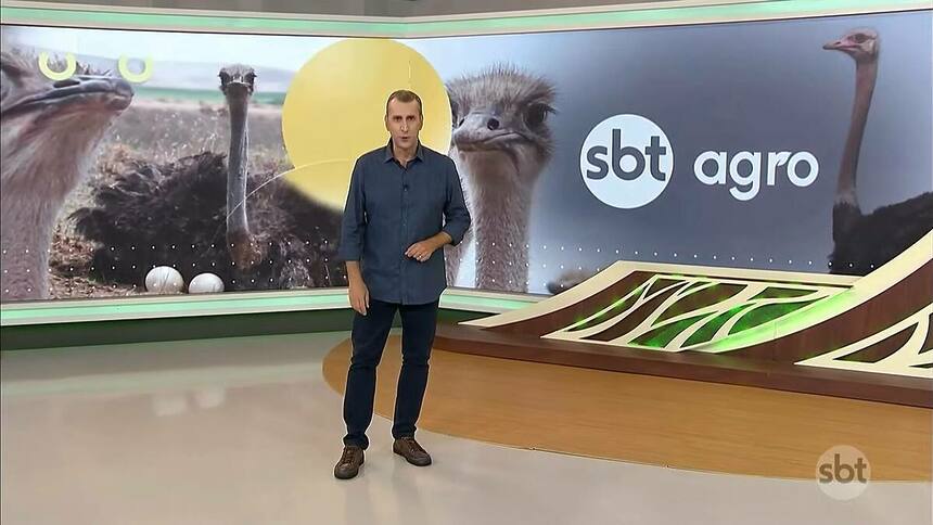 Sandro Ivanowski de camisa azul e calça azul mais escura, no centro do cenário do ABT Agro que mostra um telão com avestruzes