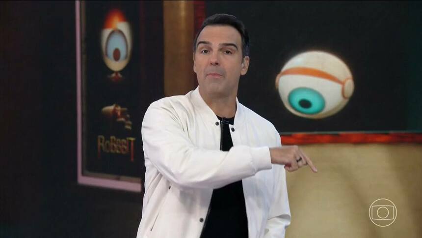 Tadeu Schmidt de camisa preta e casaco branco apontando para o logo da Globo no cenário do BBB 25