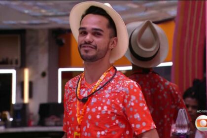 Com chapéu de sambista e camisa laranja, João Pedro sorrir enquanto distribui as pulseiras do Na Mira do Líder na sala do BBB 25