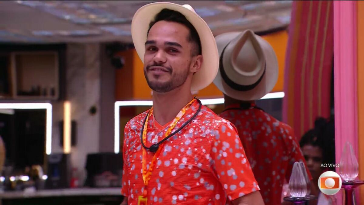 Com chapéu de sambista e camisa laranja, João Pedro sorrir enquanto distribui as pulseiras do Na Mira do Líder na sala do BBB 25