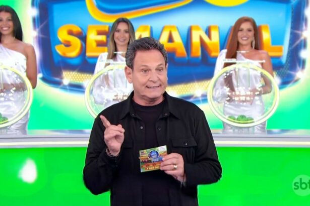 Luis Ricardo levanta a mão para iniciar o sorteio da Tele Sena Semanla edição 50. O cenário está verde e e ele se posiciona a frente dos globos com as bolas do sorteio