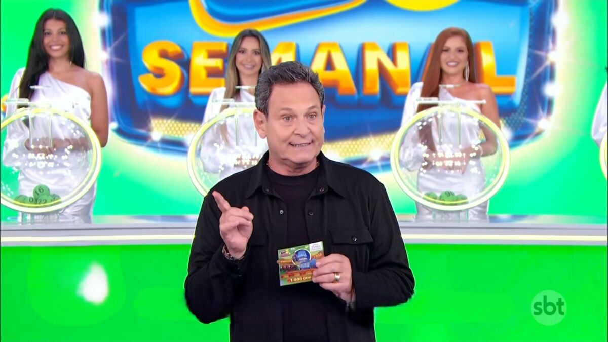 Luis Ricardo levanta a mão para iniciar o sorteio da Tele Sena Semanla edição 50. O cenário está verde e e ele se posiciona a frente dos globos com as bolas do sorteio