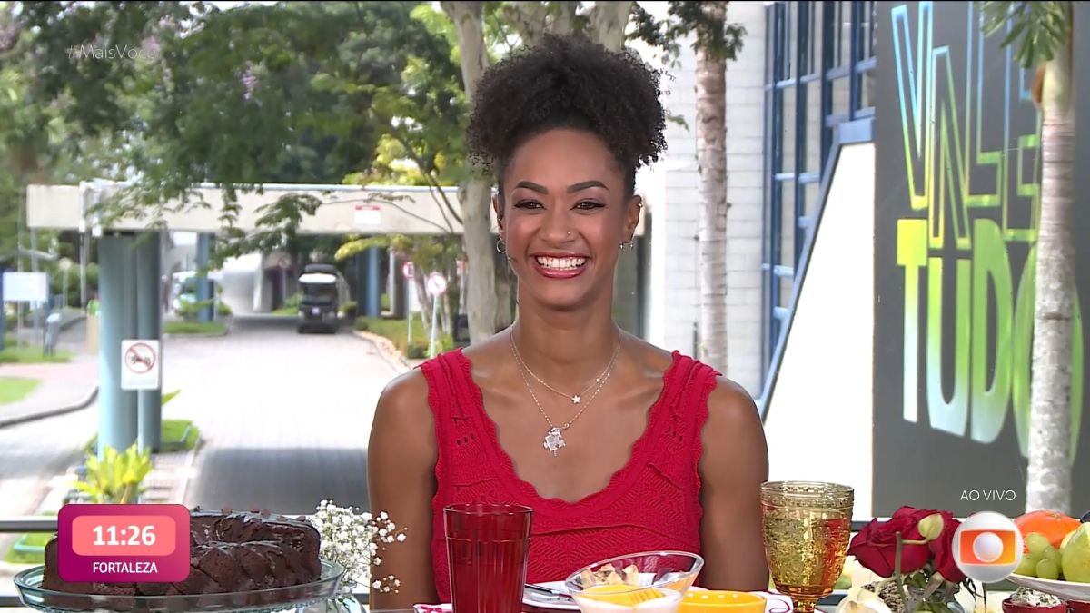 Aline de vestido vermelho, sentada na cadeira do estúdio de vidro do Mais Você no Rio de Janeiro. Ela está sorrindo