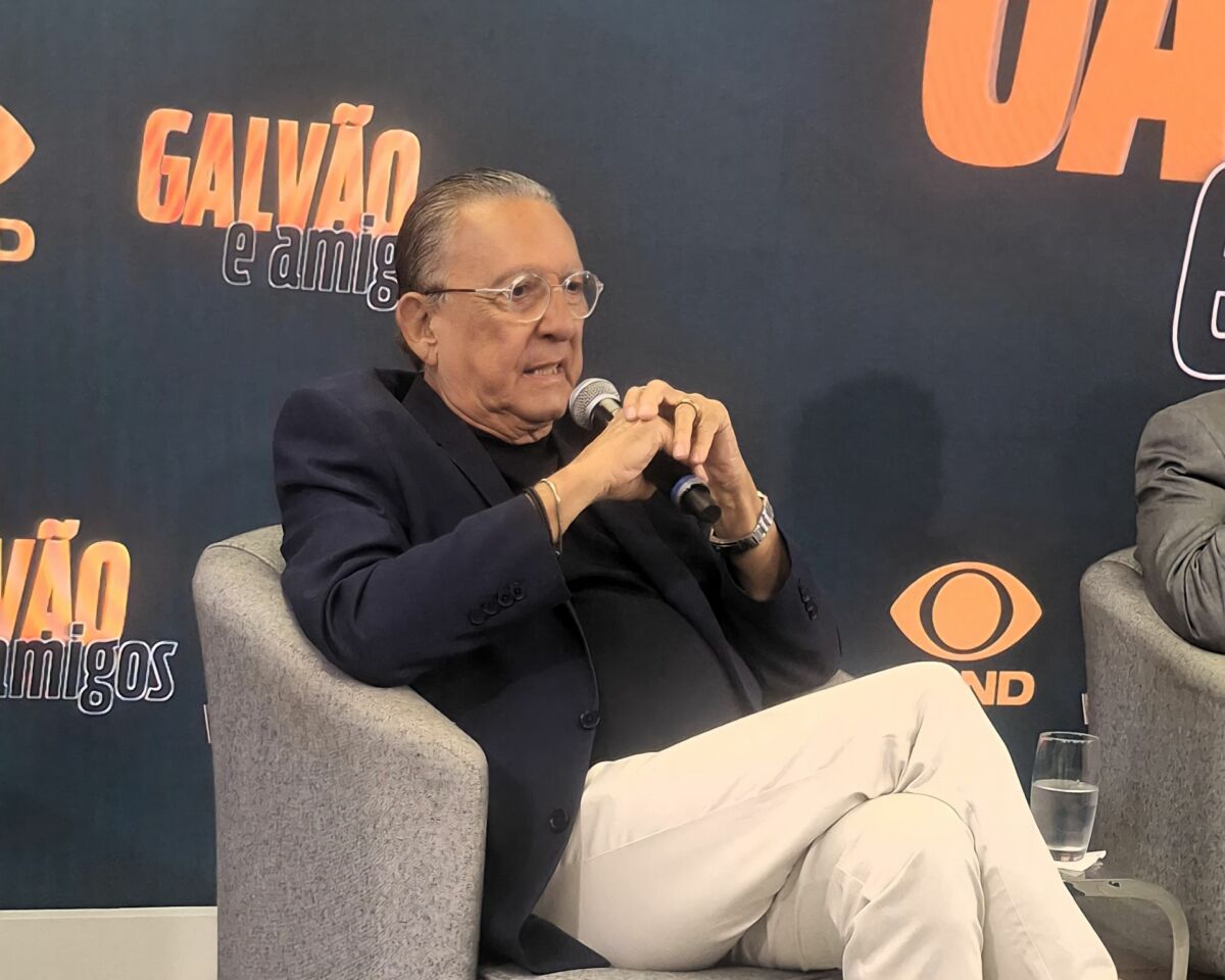 Galvão Bueno de terno preto e calça clara sentado na poltrona da Band em frente ao telão com o logo do seu novo programa Galvão e Amigos