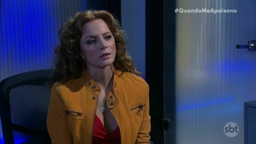 Silvia Navarro como Renata, de Quando Me Apaixono. Ela está de jaqueta marrom sentada em uma cadeira de escritório