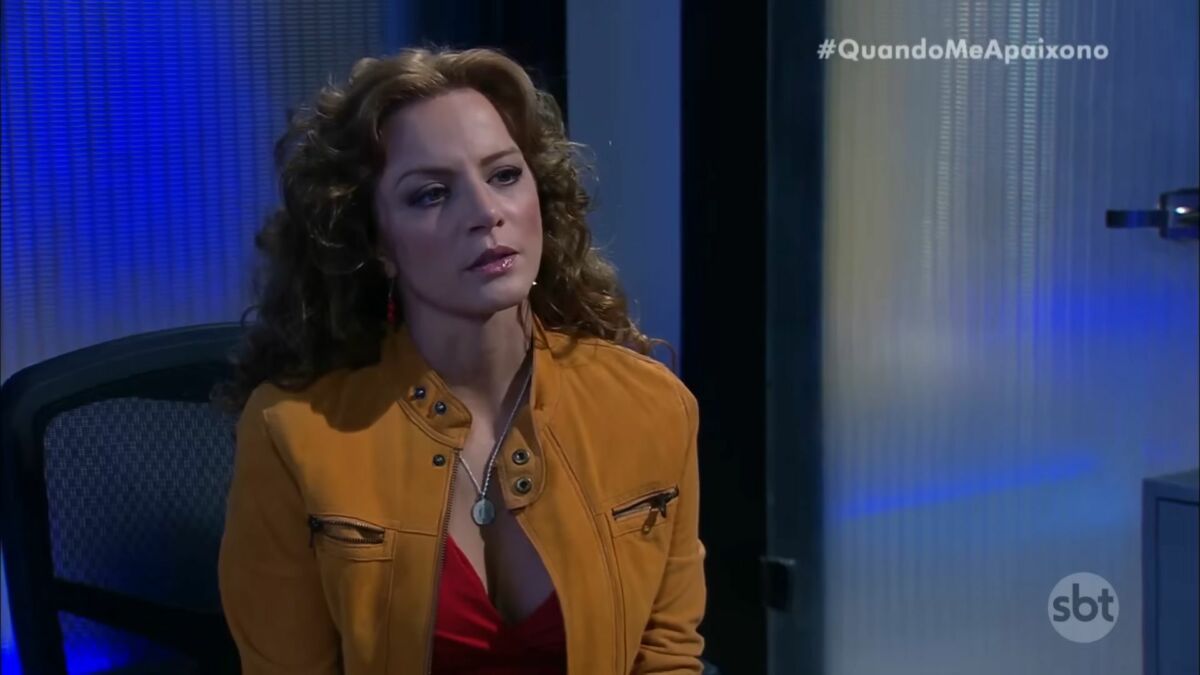 Silvia Navarro como Renata, de Quando Me Apaixono. Ela está de jaqueta marrom sentada em uma cadeira de escritório