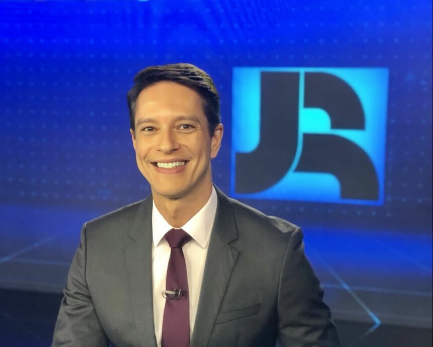 Sergio Aguiar sorridente no cenário do Jornal da Record em frente ao logo do telejornal