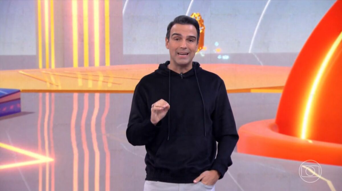 Tadeu Schmidt de macacão preto no cenário do BBB 25