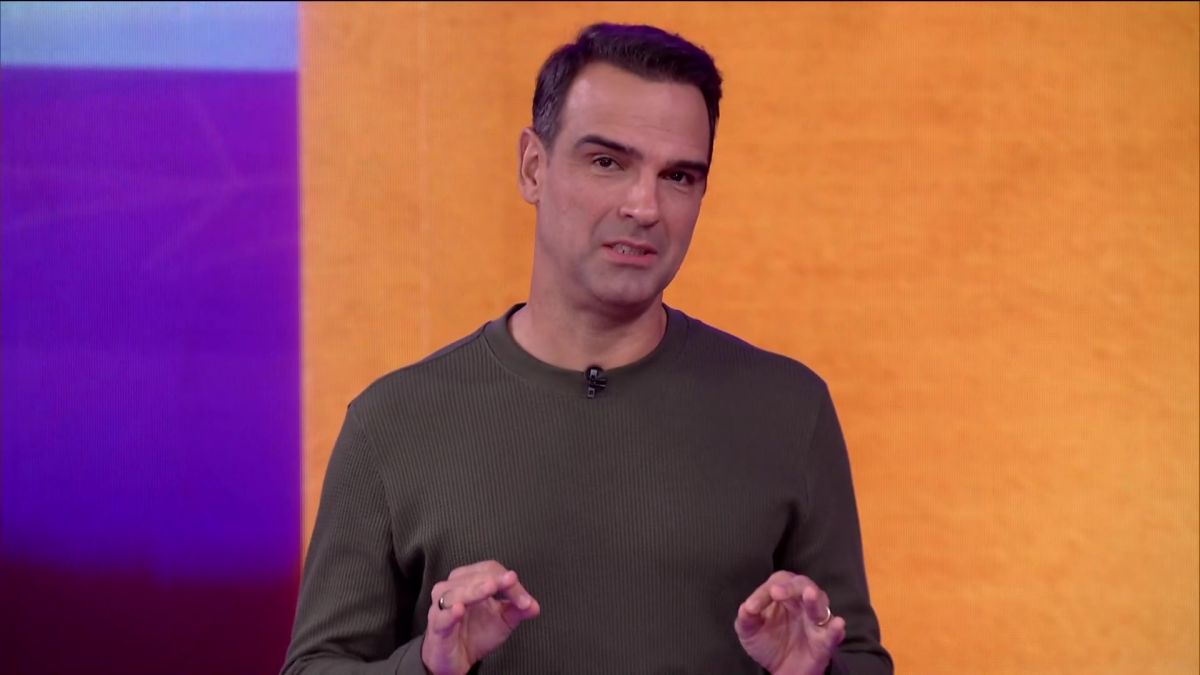 Tadeu Schmidt com camisa cinza e as mãos levantadas no cenário do BBB 25