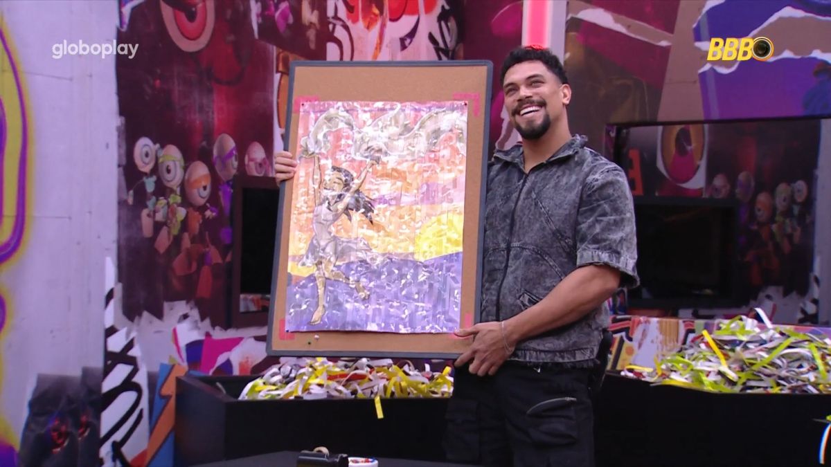 Vinicius segura quadro de fitas com desenho de Renata no quarto Barrado no Baile do BBB 25. O brother sorrir para a câmera