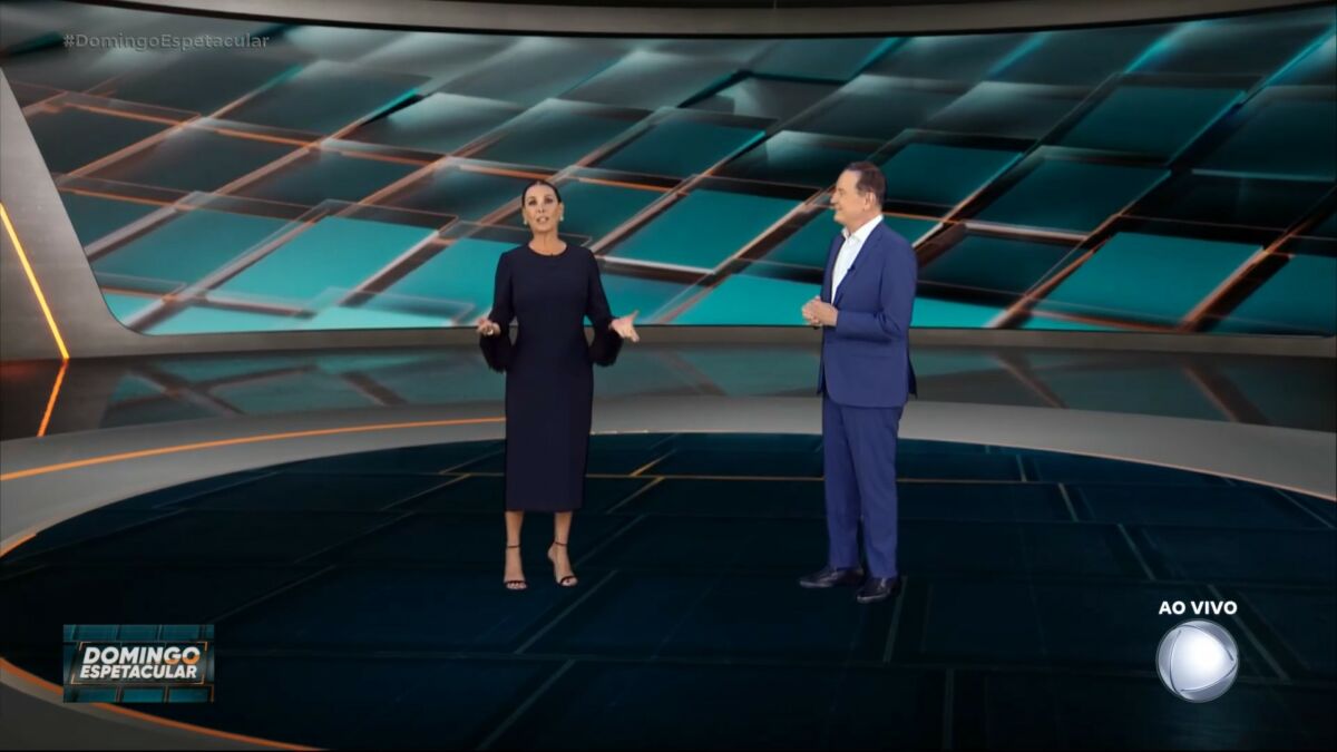 Carolina Ferraz, de vestido preto e com as mãos abertas ao lado de Roberto Cabrini na apresentação do Domingo Espetacular