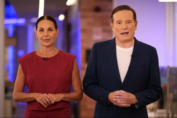 Carolina Ferraz veste conjunto vermelho ao lado de Roberto Cabrini que está de terno azul e camisa branca na redação do Domingo Espetacular