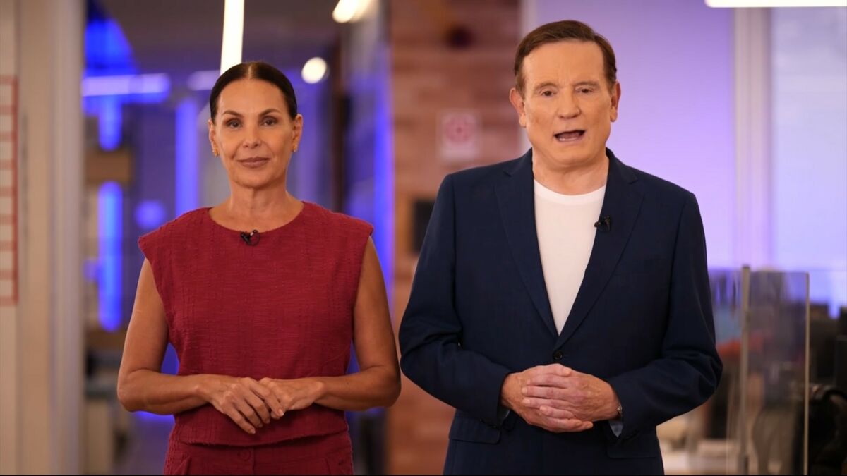 Carolina Ferraz veste conjunto vermelho ao lado de Roberto Cabrini que está de terno azul e camisa branca na redação do Domingo Espetacular