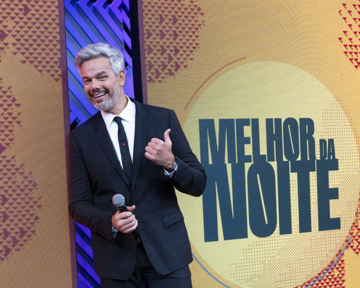 Otaviano Costa sorridente e apontando para o logo do Melhor da Noite na Band