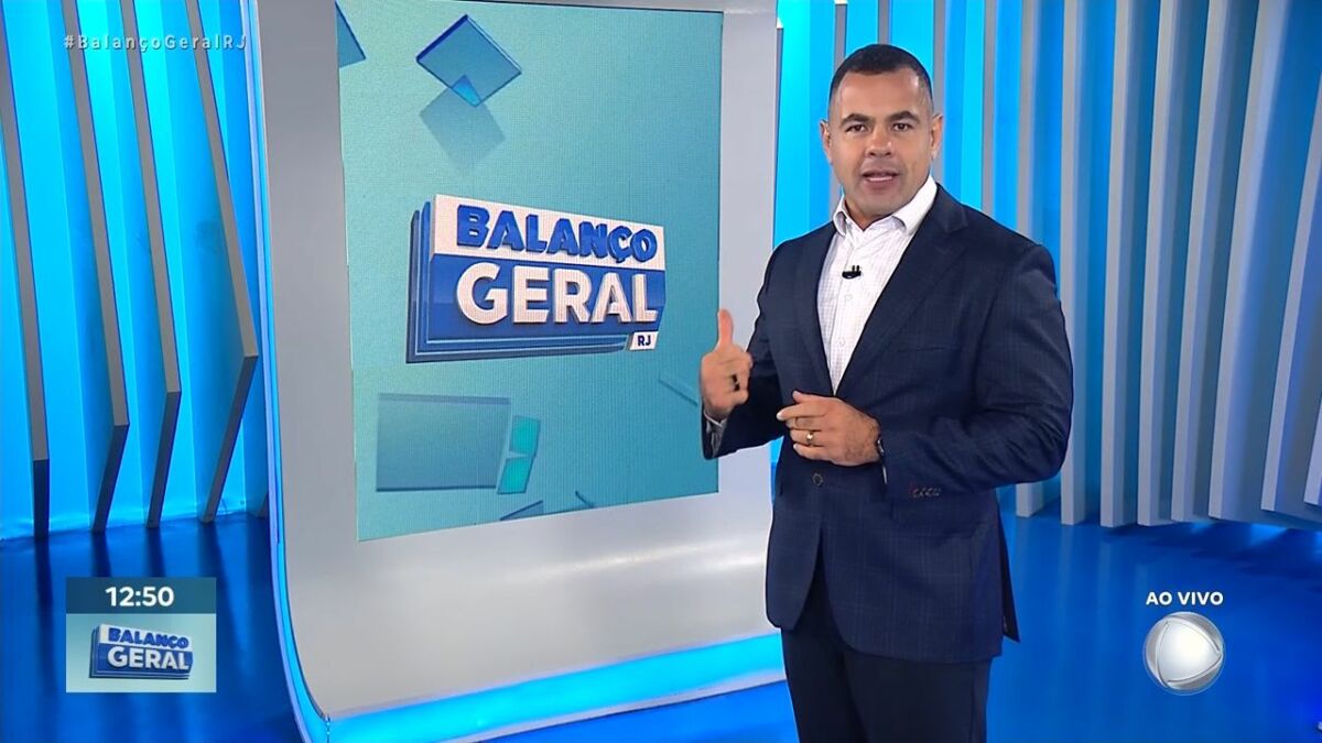 Tino Junior, de terno preto e camisa azul, fazendo sinal de legal, em frente ao telão do Balanço Geral RJ na RECORD Rio