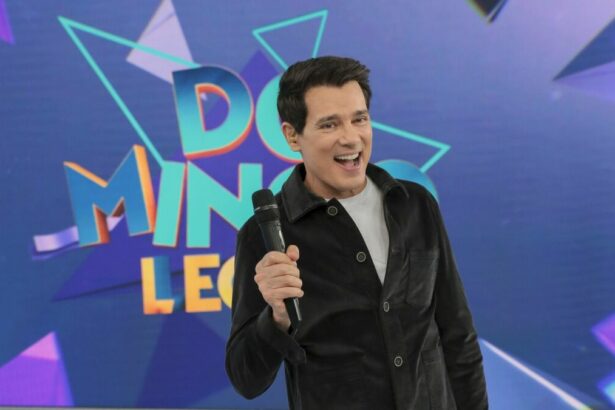 Celso Portiolli levanta o microfone e sorrir em frente ao telão do Domingo Legal