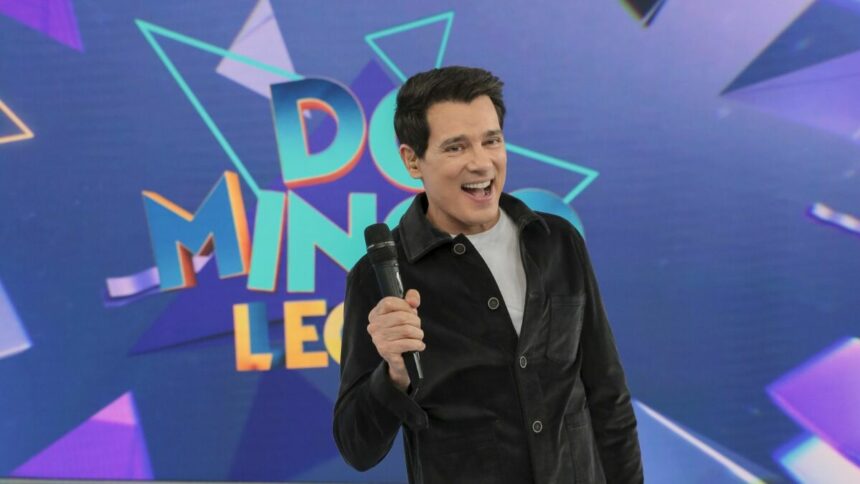 Celso Portiolli levanta o microfone e sorrir em frente ao telão do Domingo Legal