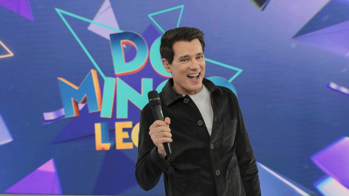 Celso Portiolli levanta o microfone e sorrir em frente ao telão do Domingo Legal