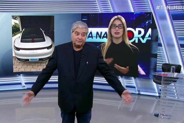 Datena abre os braços no estúdio do Tá na Hora visivelmente irritado com a repórter Nicole que aparece atrás dele no telão do SBT
