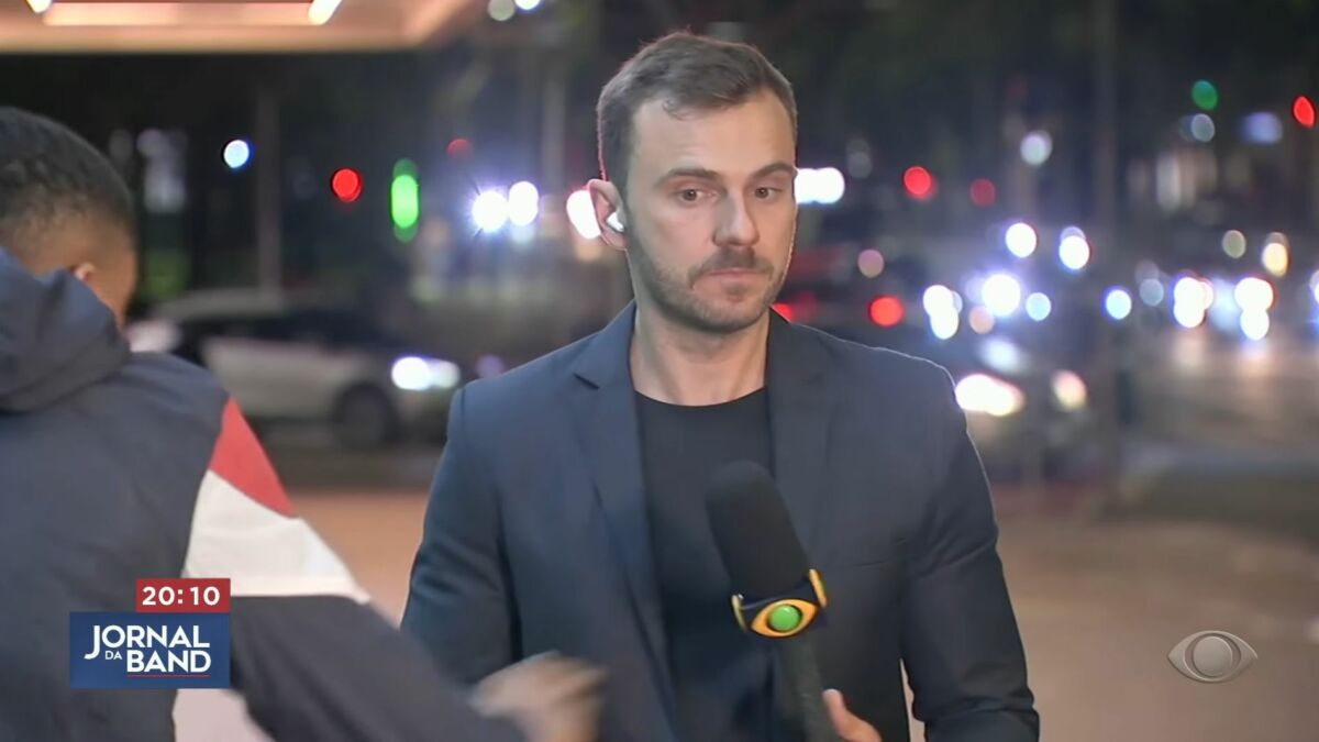 Igor Calian, com o microfone da Band nas mãos, com o ciclista passando e tentando puxar o celular
