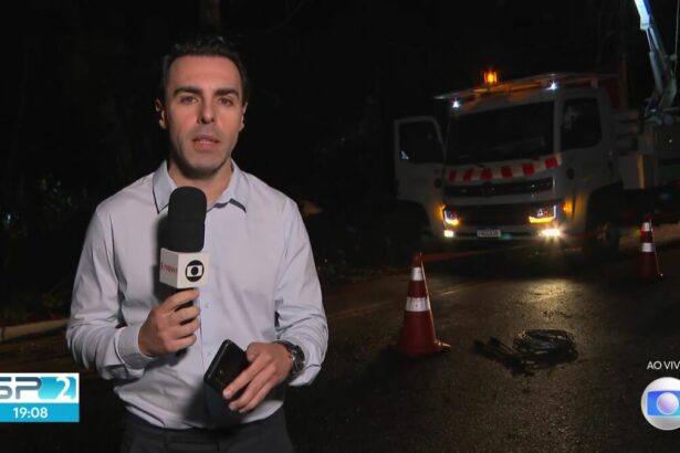 Rafael Colombo de camisa social cinza claro segurando o microfone da TV Globo em um link ao vivo em uma rua sem iluminação pública com um caminhão da companhia de energia atrás do repórter