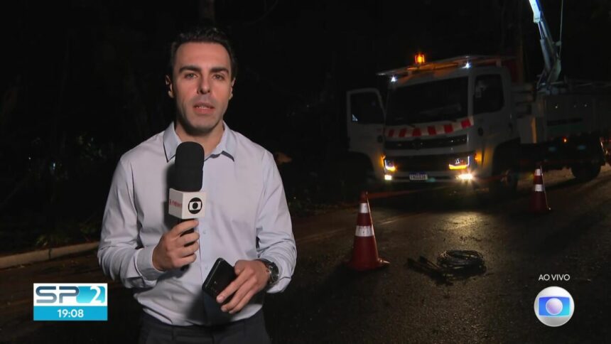 Rafael Colombo de camisa social cinza claro segurando o microfone da TV Globo em um link ao vivo em uma rua sem iluminação pública com um caminhão da companhia de energia atrás do repórter