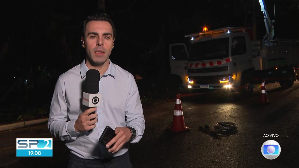 Rafael Colombo de camisa social cinza claro segurando o microfone da TV Globo em um link ao vivo em uma rua sem iluminação pública com um caminhão da companhia de energia atrás do repórter