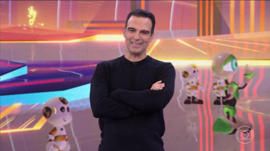 Tadeu veste camisa preta e sorrir em frente aos dummies no cenário do BBB 25