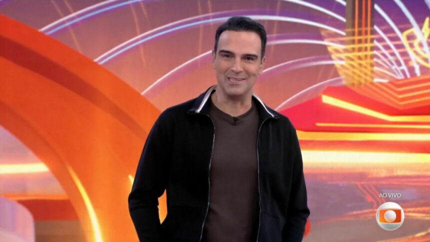 Tadeu Schmidt de camisa preta de botão e camisa cinza por dentro apresentando o BBB 25. Ele está sorrindo ao anunciar o BBB 26