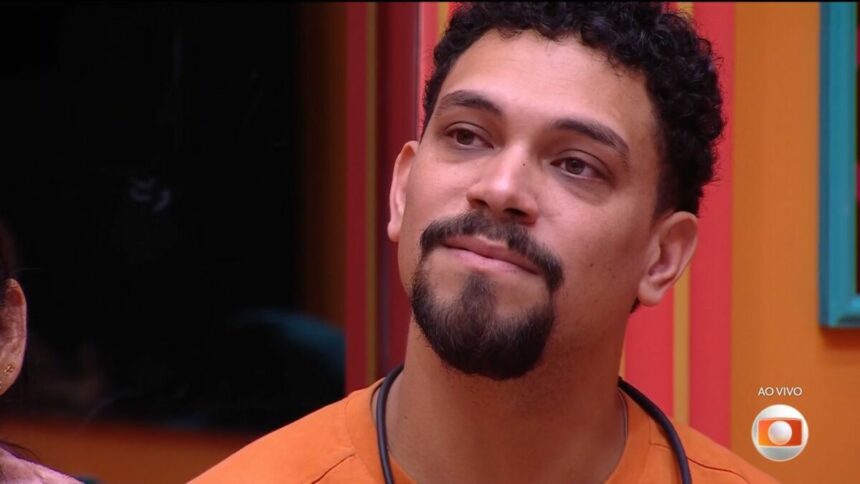 Vinicius, de camisa laranja, chora com discurso de Tadeu Schmidt antes da eliminação. Ele está sentado no sofá do BBB 25