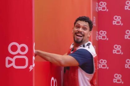 Vinicius sorrir ao lado de uma parede com o logo do patrocinador da última Prova de Anjo do BBB 25