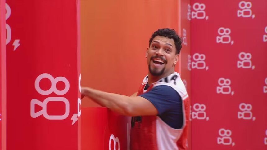 Vinicius sorrir ao lado de uma parede com o logo do patrocinador da última Prova de Anjo do BBB 25