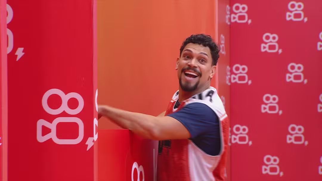 Vinicius sorrir ao lado de uma parede com o logo do patrocinador da última Prova de Anjo do BBB 25
